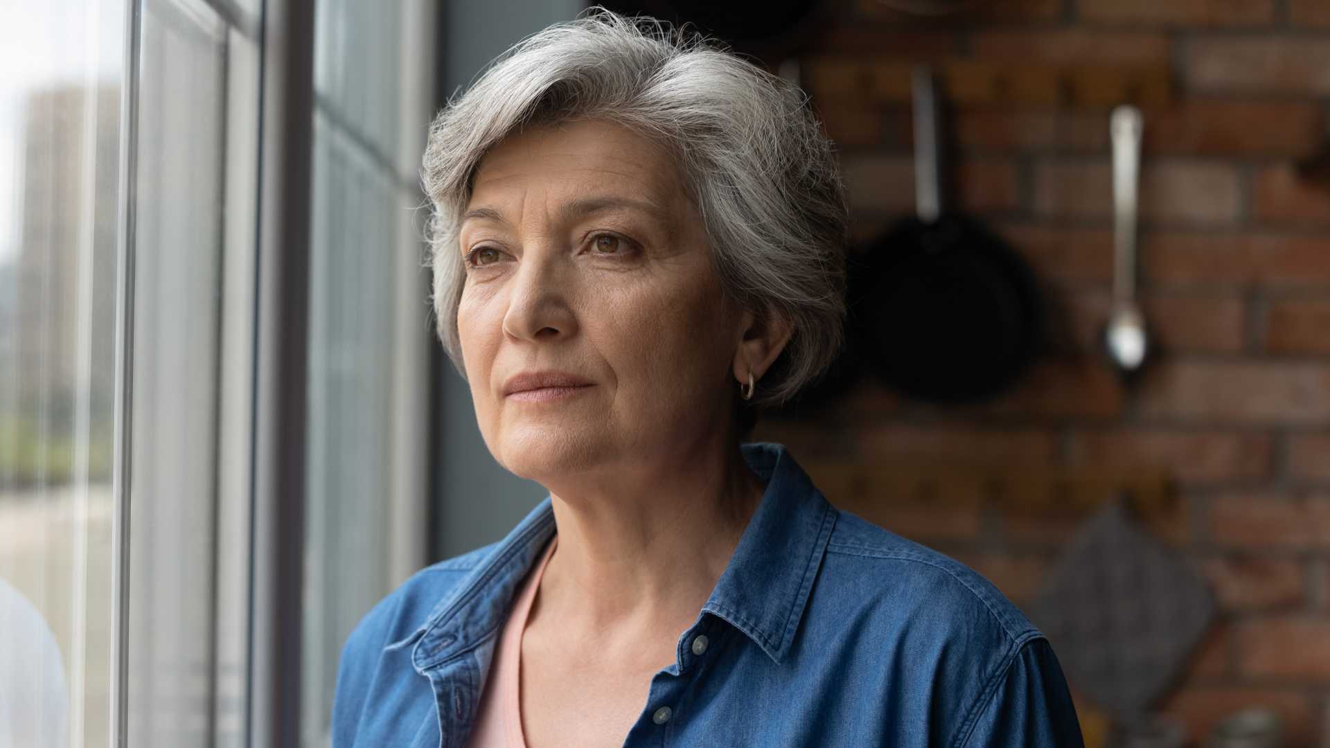 Older woman looking out window with solemn expression