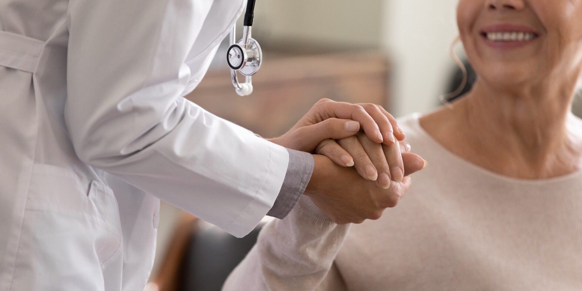 Doctor giving hope. Close up shot of young female physician leaning forward to smiling elderly lady patient holding her hand in palms. Woman caretaker in white coat supporting encouraging old person